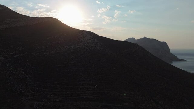 Drone trucking pan as sunlight reveals behind backlit mountains, Anafi island coastline with prominent monolith