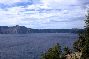 View across Crater Lake to the far lip of the crater rising in the background, sunlight sparkling on the pure blue water surface with tiny clouds overhead and tall pine trees in the foreground. 