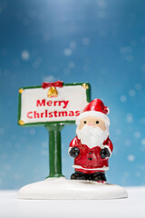 Miniature Santa Claus figurine standing next to a festive Merry Christmas sign with a snowflake-filled blue background, perfect for holiday-themed designs or decorations.