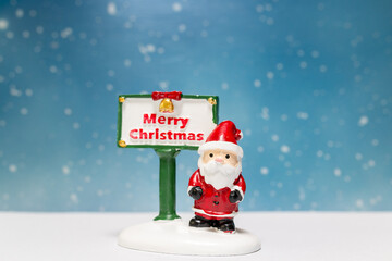 Miniature Santa Claus figurine standing next to a festive Merry Christmas sign with a snowflake-filled blue background, perfect for holiday-themed designs or decorations.