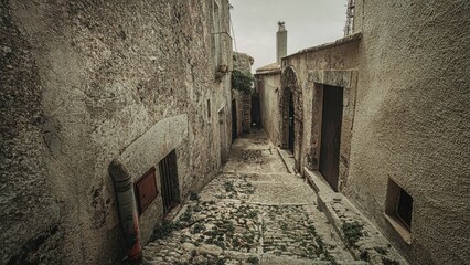Narrow cobblestone alley in Erice, Sicily, Italy with rustic stone walls and a cloudy sky