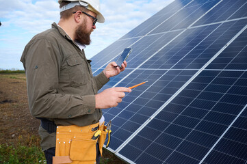 Engineer in uniform with protective helmet near solar station holding screwdriver. Supervisor is checking on solar panels on solar farm. Green energy concept.