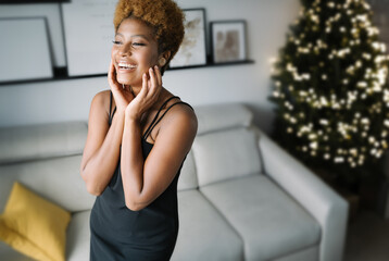 Fototapeta premium Happy laughing black woman in elegant little dress in cozy light interior decorated Christmas tree. Winter holidays magic moment. Closing eyes and touching her face with her hands. Wow emotions
