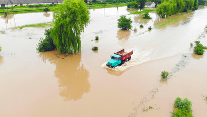 Truck driving through flooded area after heavy rainfall. A truck navigates through deep floodwaters covering a roadway, surrounded by partially submerged trees and vegetation.