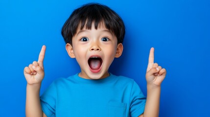 Young boy is standing in front of a blue wall with his hands raised and pointing upwards. He is smiling and he is excited or happy