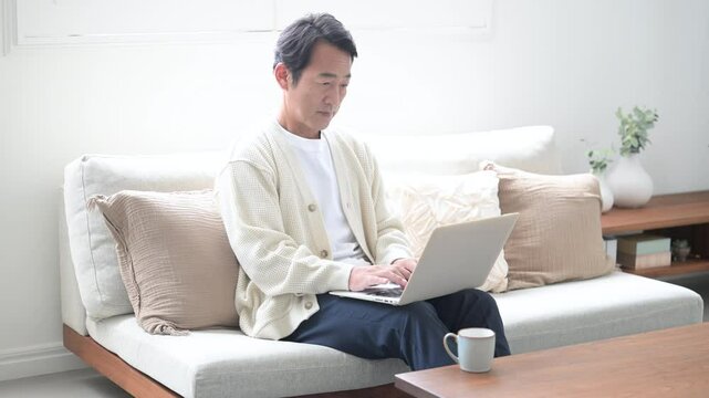 綺麗なリビングでノートパソコンを使う、お茶を飲む長袖のカーディガンを着た50代の男性の動画