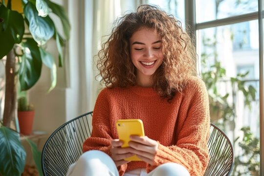 Young Woman Smiling Sitting on Wire Chair Holding Smartphone Using Mobile Apps Relaxing in Bright Room with Plants