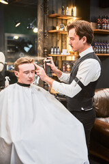 Professional barber using hairspray to finish hairstyle. Stylish hairdresser making final touches. Young customer getting fresh haircut.