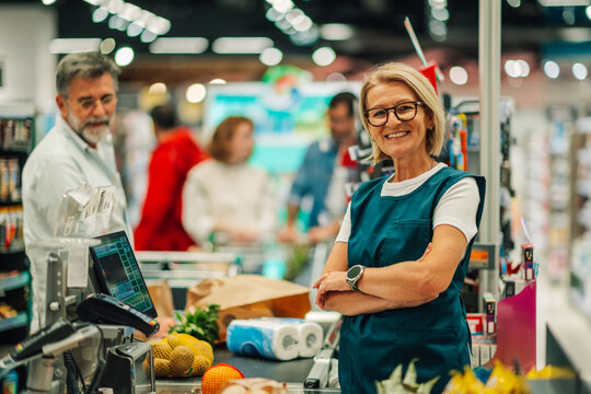 Supermarket cashier smiling at checkout counter with customers waiting