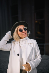 A woman in a stylish white coat and hat enjoys a sunny day in an urban setting