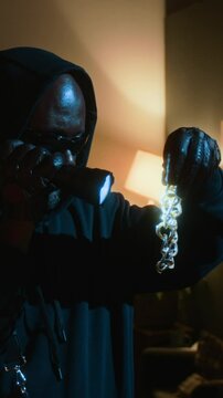 Vertical shot of male thief in black sweatshirt with glasses shining flashlight at golden chain while carrying out burglary in apartment