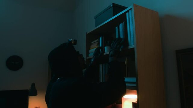 African American criminal checking books, then taking box off bookcase, opening, rummaging through it and finding rolls of banknotes while pulling heist