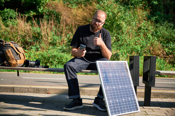 Bearded man using smartphone connected to photovoltaic solar panel, giving thumbs up. Integration...