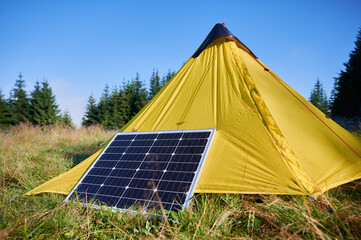 Yellow tent with solar panel set up in grassy field surrounded by dense evergreen trees under clear...