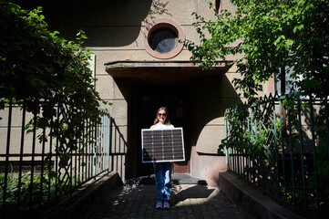 Woman holding solar panel for charging, stands in front of old building's entrance. Smiling girl...