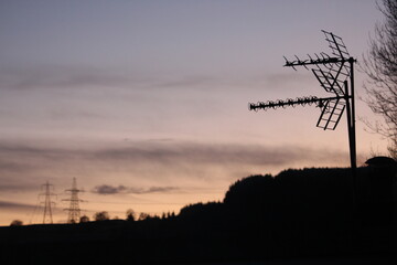 A photograph of a tv aerial on a roof with electricity pylons in the background.  Silhouette against sunset sky