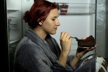 Beautiful young woman eating unhealthy food near refrigerator at night