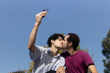 Gay couple kissing while taking a selfie outdoors. copy space