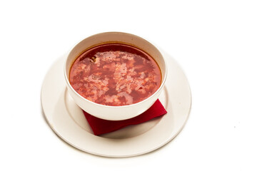 A bowl of red soup sits on a white plate with a red napkin underneath