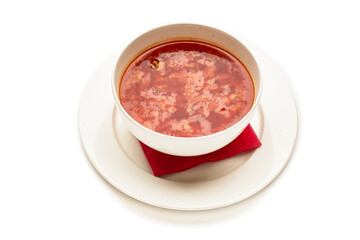 A bowl of red soup sits on a white plate with a red napkin underneath