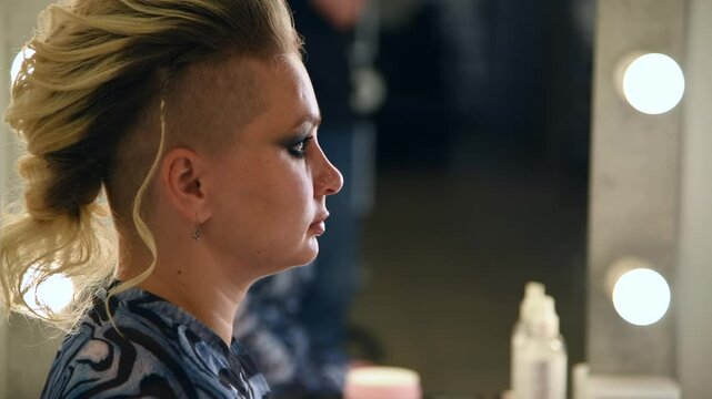 Side view of make-up artist applying eyeliner on eyes of adult blond haired woman with mohawk hairstyle in beauty salon. Soft focus. Real time handheld video. Makeup and beauty industry theme.