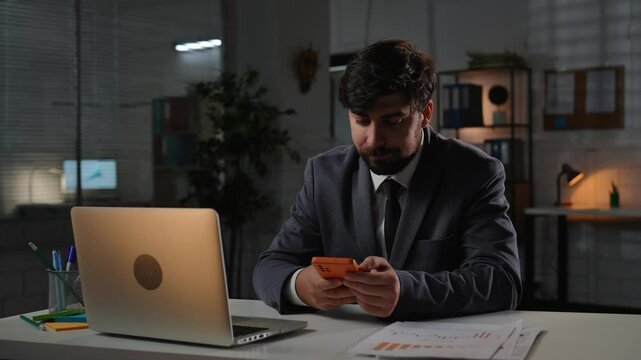 Adult business man manager sitting at desk working on laptop in office overtime in evening, having break watching social media on smartphone.
