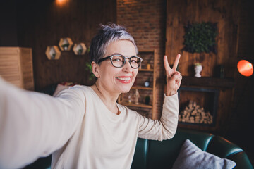 Happy senior woman taking a joyful selfie at home, with a charming smile and peace sign, enjoying...