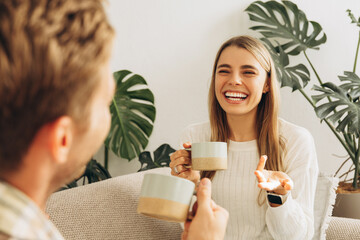 Beautiful couple holding cup with coffee, sitting on comfortable sofa, talking each other at home