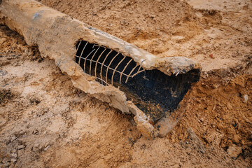 A heavily corroded pipe with exposed metal reinforcement lies damaged in the dirt, illustrating...