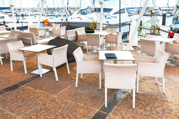 Outdoor seating area at a marina, with tables and chairs arranged on a tiled floor. In the background, there are boats docked at the marina, adding a scenic and nautical atmosphere to the setting