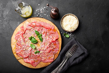 Slices of fresh beef carpaccio with basil and parmesan cheese on a wooden cutting board on the black background top view