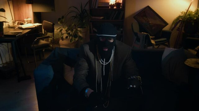 Portrait of brutal Caucasian young male thief wearing balaclava and necklaces holding crowbar and stroking his chin while sitting on sofa in messy apartment
