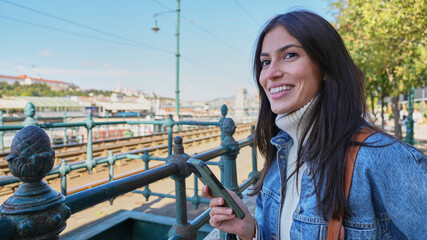 A casually dressed woman walks through a park in the city, scrolling through her phone with a smile.