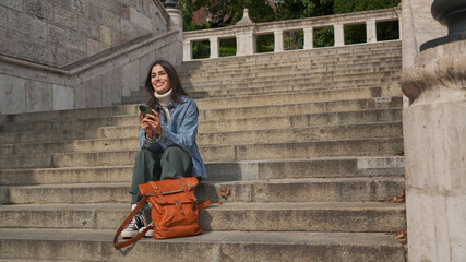 Beautiful young smiling woman sitting on steps and reading text messages from friends on a...