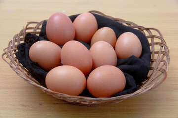 A basket of eggs sits on a wooden table. The eggs are brown and appear to be fresh. The basket is...