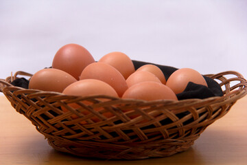 A basket of eggs sits on a wooden table. The eggs are brown and appear to be fresh. The basket is...