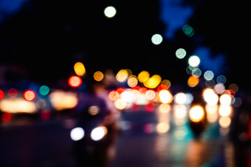 A blurry image of a busy street at night with cars, motorcycles, and pedestrians. The scene is filled with bright lights and the overall mood is energetic and bustling
