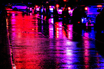 A group of people are standing on a wet street at night. The street is illuminated by the lights of cars and police cars. The scene is dark and moody © SAKCHAI