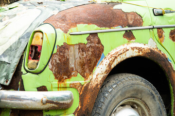 A green car with rusted parts and a broken headlight. The car is old and has been left to rust