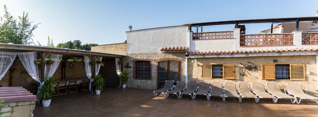 Naklejka premium Sunlit patio of a mediterranean villa featuring a canopy with white curtains, a dining area, and several deck chairs for relaxation