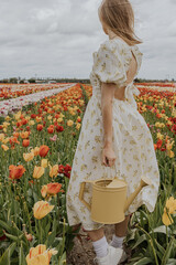 A girl in a yellow dress and apron with a water can in a red and green tulip flower field on a gloomy rainy cloudy sky background happily running around. 

