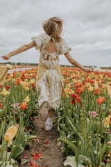 A girl in a yellow dress and apron with a water can in a red and green tulip flower field on a gloomy rainy cloudy sky background happily running around. 

