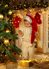 Christmas Child with Xmas Golden Ornament in Cozy Room with Fireplace. Baby in Santa Hat next to Christmas Tree decorated with Gold Toys and Lights Garland