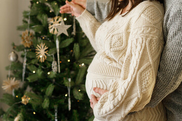 Happy pregnant couple hugging belly and decorating together christmas tree. Family bonding and love concept. Pregnancy during holidays, magical joyful time.