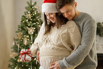 Happy pregnant christmas family hugging belly and holding together christmas gift at festive...