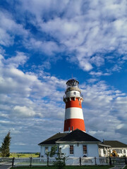 A striking white lighthouse stands amid a field, under a blue sky scattered with clouds. The pastoral setting evokes calmness, suitable for travel, rural lifestyle, or coastal themes.