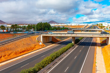 Highway in Arrecife, Lanzarote, with an overpass bridge crossing over it. Highway is divided by a median strip with greenery, there are multiple lanes in each direction. Overpass road with vehicles