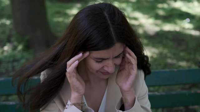Businesswoman sitting on a bench, struggling with a headache while surrounded by a park's calming nature in slow motion