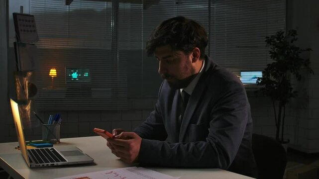 Adult business man manager sitting at desk working on laptop in office at night, having break watching social media on smartphone, close up.