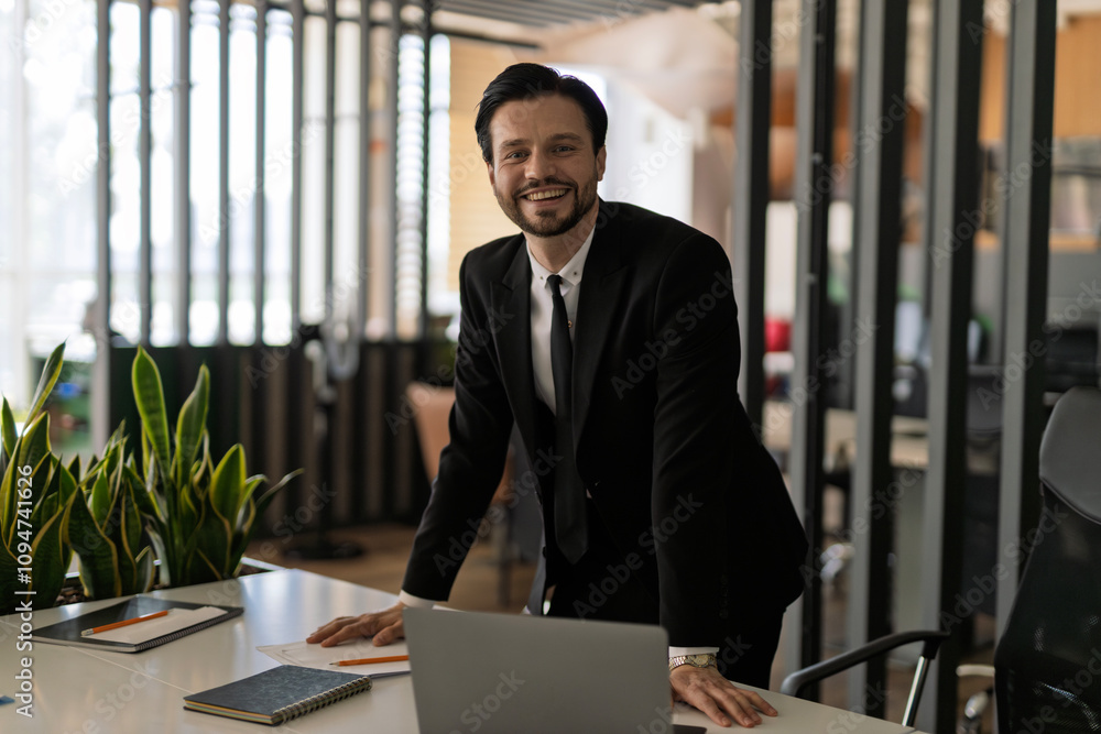 Poster a man in a suit stands in front of a desk with a laptop and a potted plant. he is smiling and he is 